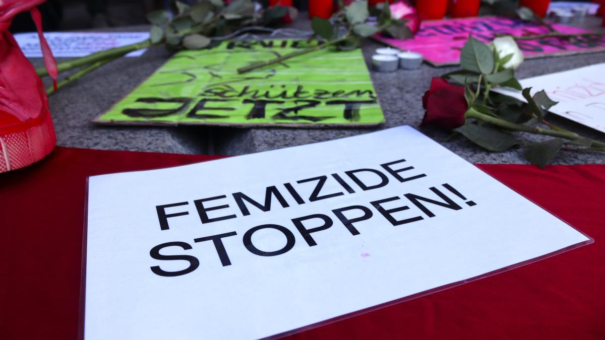 Auf einem Plakat ist zu lesen: Femizide stoppen! Dahinter sind Grabkerzen und Blumen zu sehen.