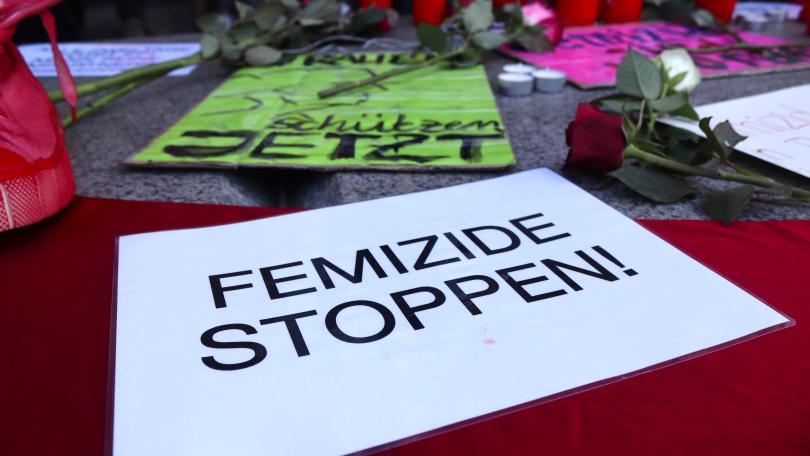 Auf einem Plakat ist zu lesen: Femizide stoppen! Dahinter sind Grabkerzen und Blumen zu sehen.